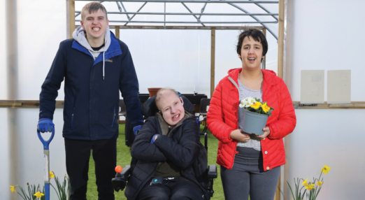 Two male students and female student standing in greenhouse holding gardening tools and plants