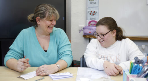 Tutor wearing a turquoise jumper supporting a female student as they both sit behind a desk