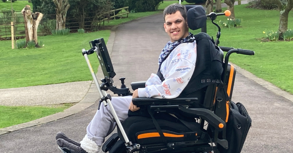 Laith sitting in his wheelchair smiling with the National Star campus in the background